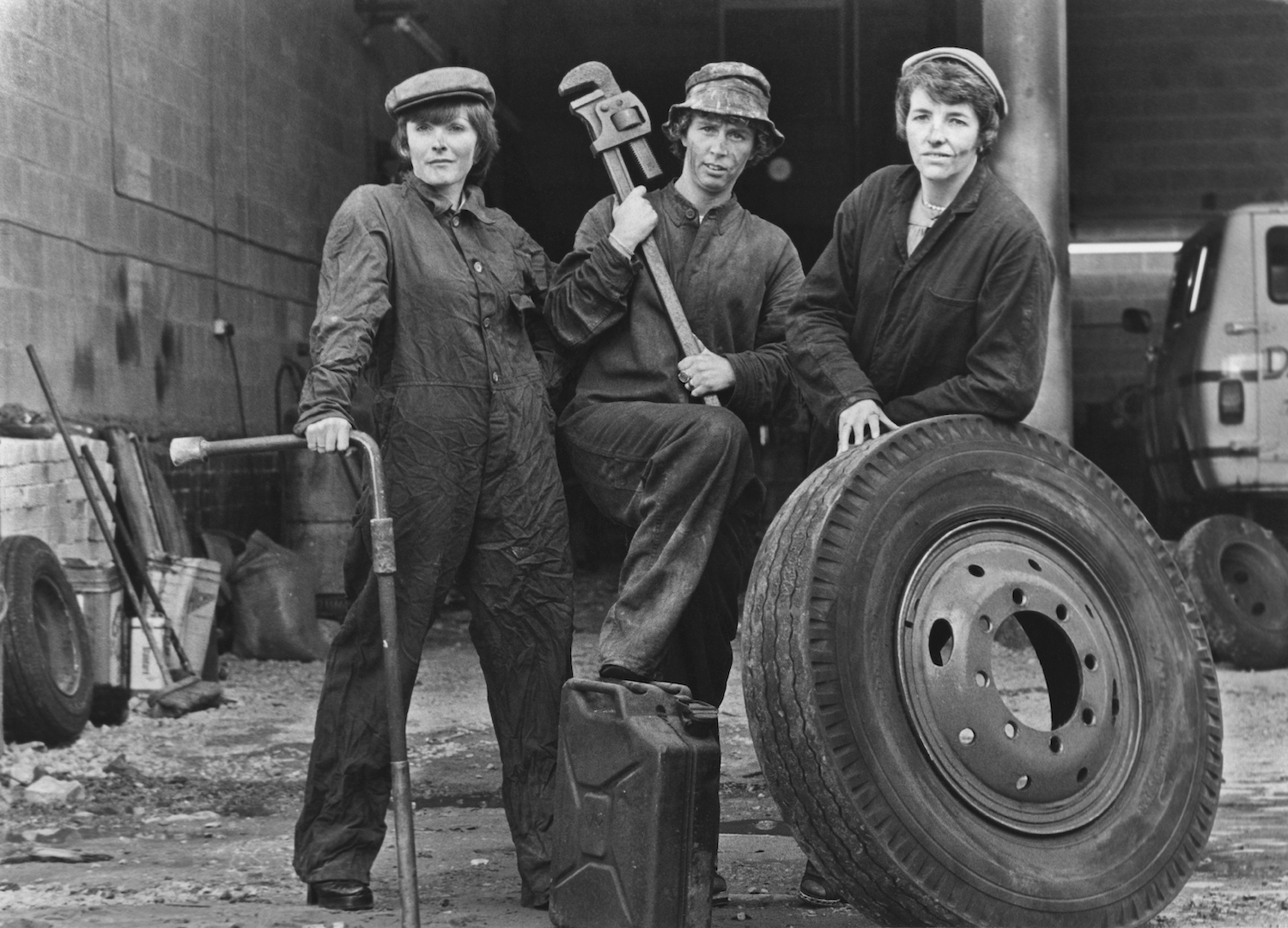 Three 1960s female mechanics pose for a black and white photo with a truck tire, a pipe wrench, and a lug wrench with a breaker bar extension to break nuts free.