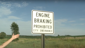 A white "Engine Braking Prohibited: City Ordinance" road sign with a grassy field visible behind it.