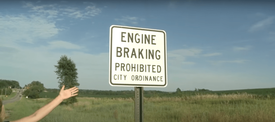 A white "Engine Braking Prohibited: City Ordinance" road sign with a grassy field visible behind it.