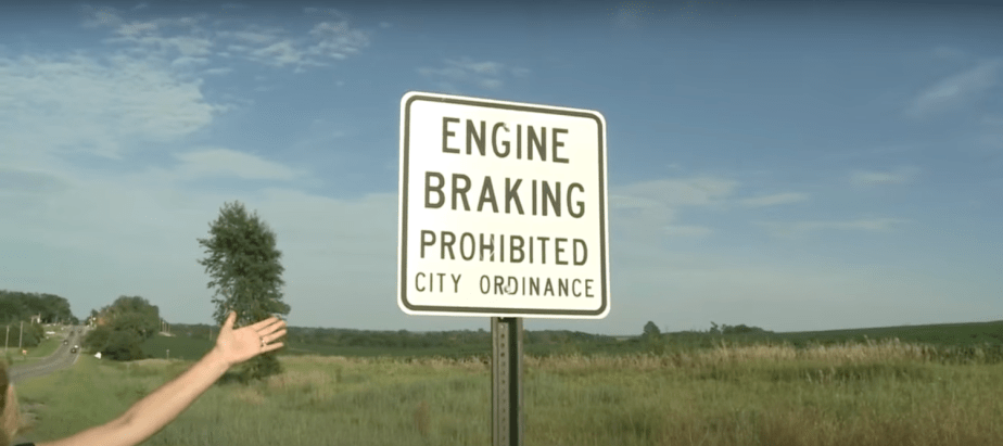 An "Engine Braking Prohibited" sign on a roadside, a grassy field visible in the background.