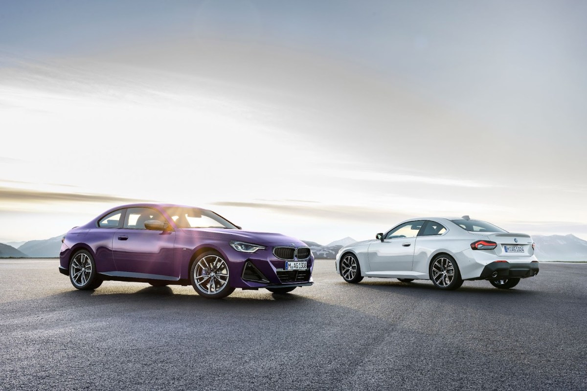 Two BMW 2 Series Coupes, one in purple on the left, one in white on the right. Parked facing each other over a mountain landscape