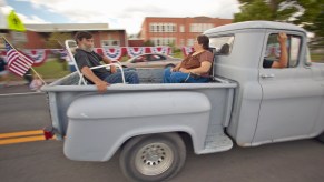 A couple rides in the bed of a primer gray stepside pickup truck during a parade.