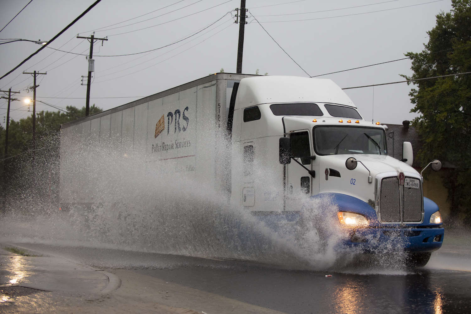 A semi truck driving through standing water and slowing down, trees visible in the background.