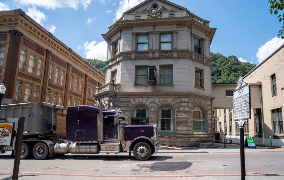 A semi truck driving through a downtown district, stone brick buildings visible in the background.