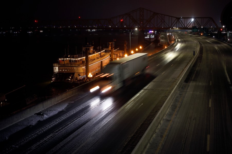 A semi truck driver flashing their high beam headlights on the interstate highway at night, a city skyline visible in the backgound.