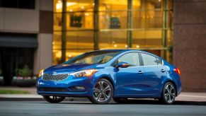 A blue 2014 Kia Forte compact sedan model parked outside of a glass-windowed office building