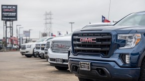 The grilles of a row of GMC and Chevrolet pickup trucks parked at a General Motors dealership, a row of buildings visible in the background.