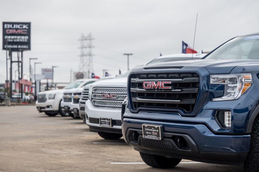 The grilles of a row of GMC and Chevrolet pickup trucks parked at a General Motors dealership, a row of buildings visible in the background.