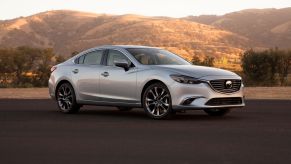 A light-gray 2016 Mazda6 midsize sedan parked on an asphalt lot near dry grass hills