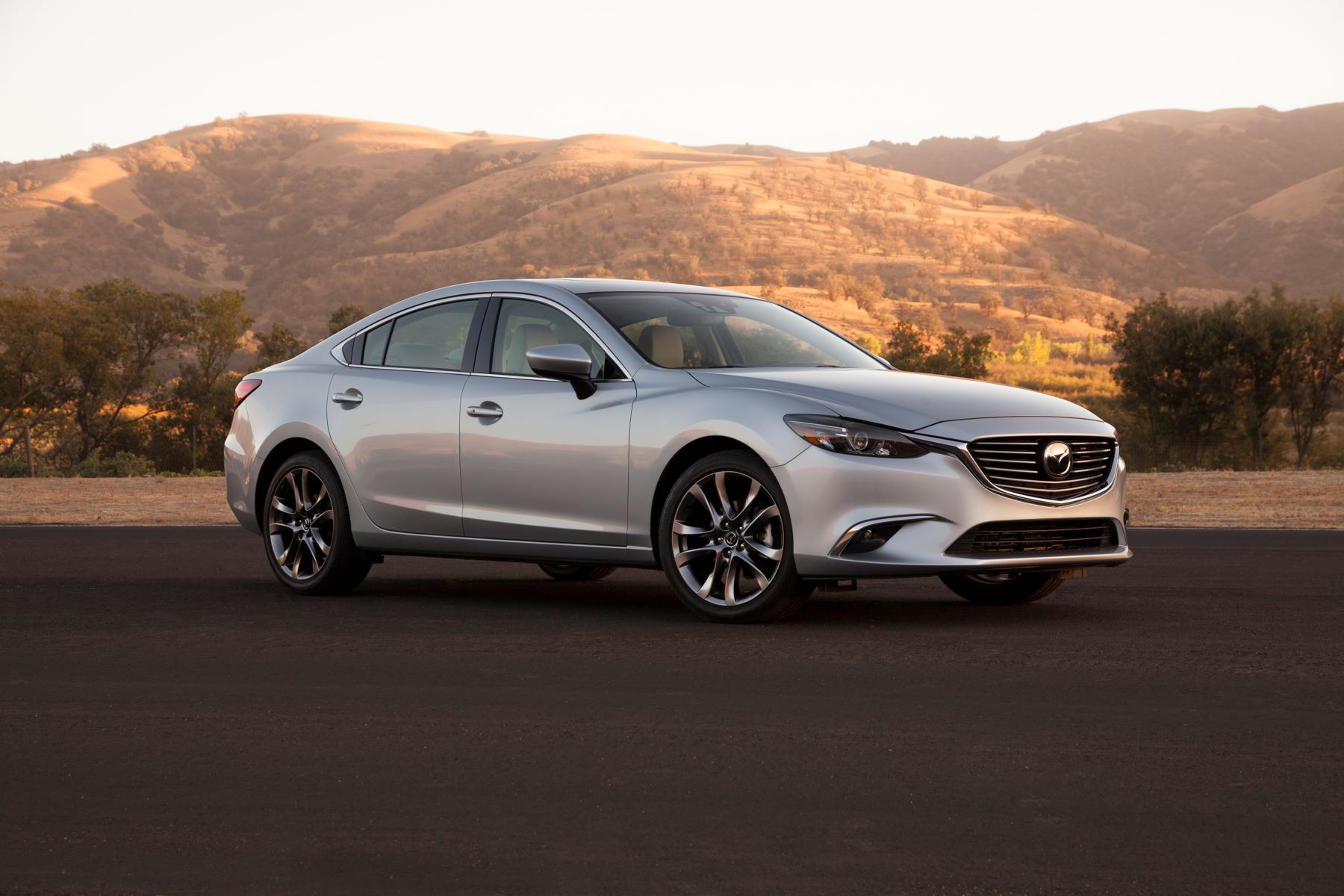 A light-gray 2016 Mazda6 midsize sedan parked on an asphalt lot near dry grass hills