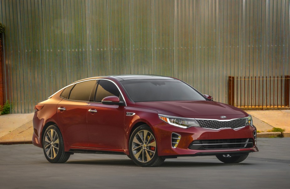 A reliable, used red Kia Optima parks next to a large building.