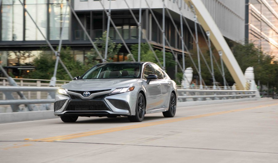 A Toyota Camry Hybrid shows off its silver paintwork while it cruises across a bridge in a city.