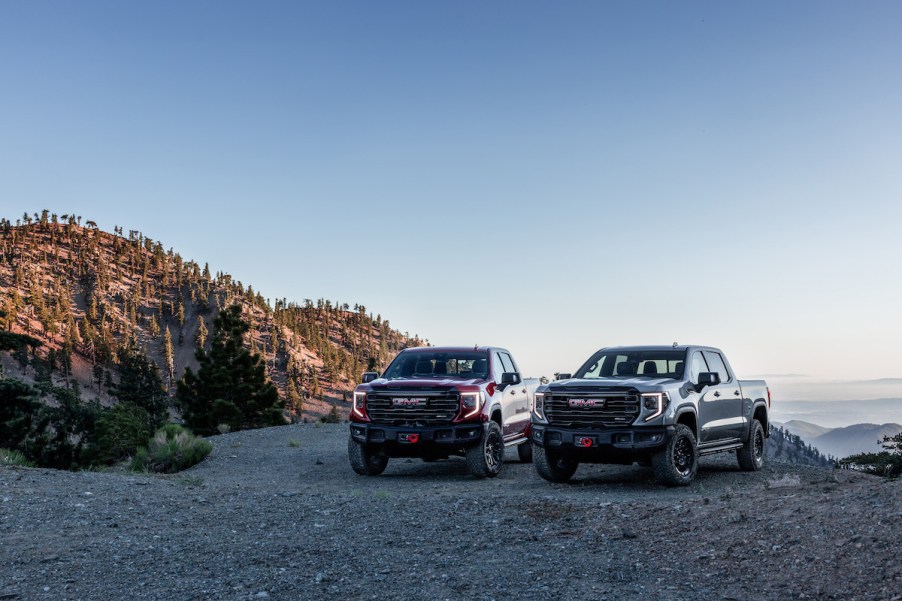 Two 2023 GMC Sierra trucks parked outdoors in front of a mountain and body of water.