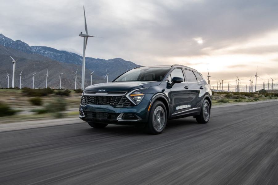 A blue-green 2023 Kia Sportage Hybrid compact SUV model driving past a field of windmills