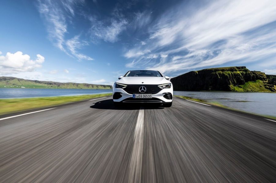 A white 2023 Mercedes-EQ EQE luxury electric car blasts down a back road along lakes.