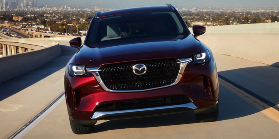 A red 2024 Mazda CX-90 midsize SUV is driving on the road. 