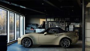 A side profile shot of a beige 2023 Mazda MX-5 Miata convertible roadster model parked inside a garage