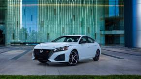 A white 2023 Nissan Altima midsize sedan model parked on an office plaza in front of glass plating