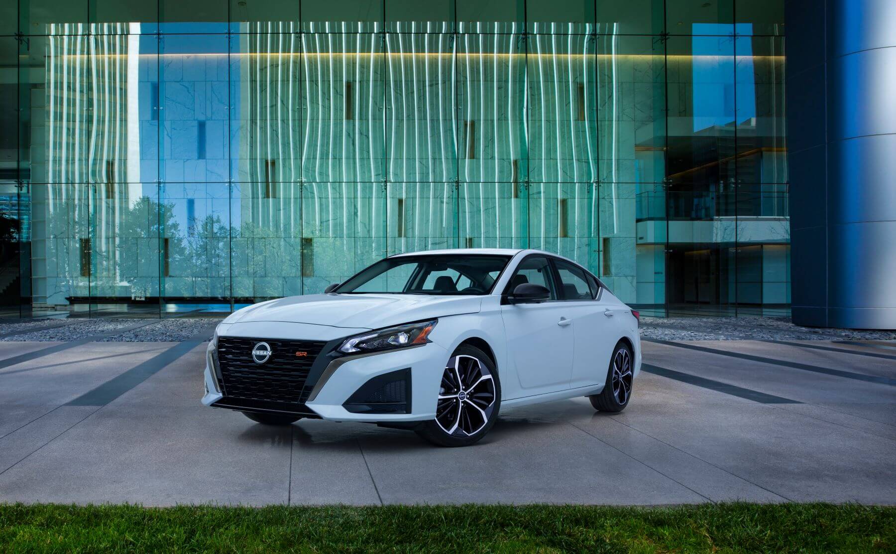 A white 2023 Nissan Altima midsize sedan model parked on an office plaza in front of glass plating