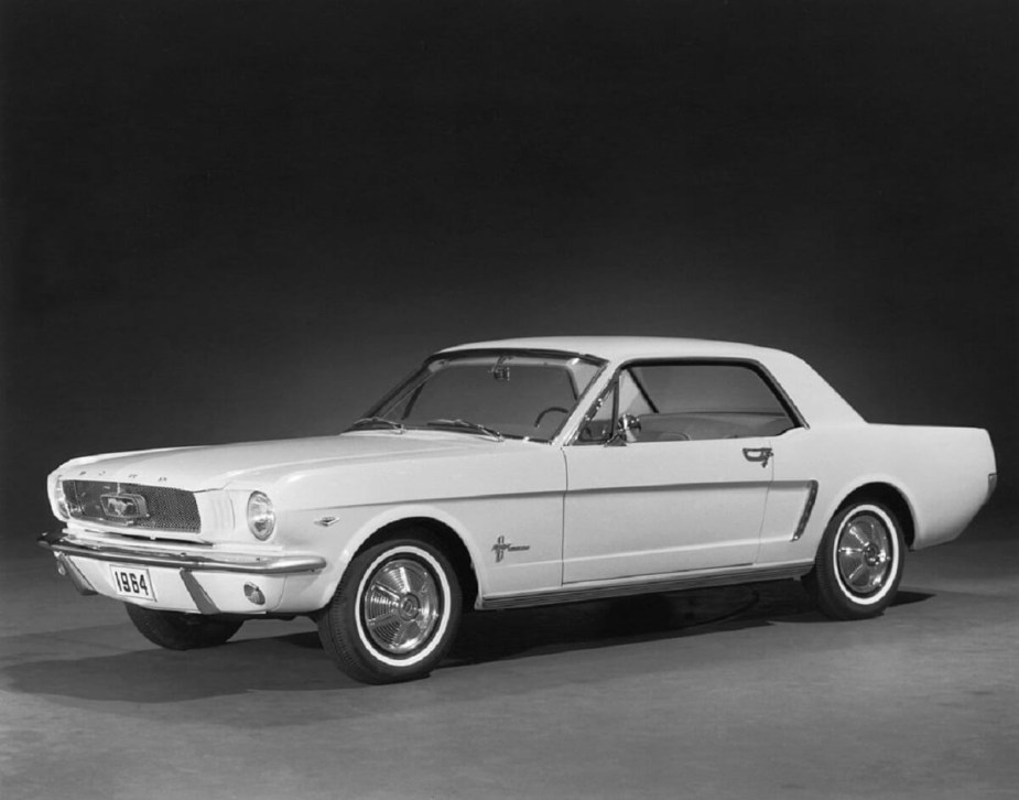 A first-generation 1965 Ford Mustang, what fans call the 1964 1/2 model, shows off its white paintwork and Ford Falcon platform on a stage.