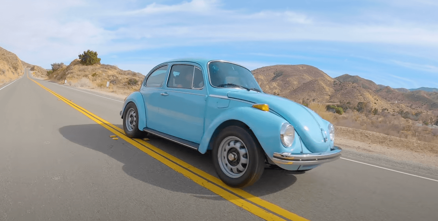 Blue 1973 Volkswagen Beetle racing along a remote road in California, desert ridgelines visible in the background.