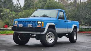 A blue 1990 Jeep Comanche Cherokee-based 4WD pickup truck in a parking lot for an auction photoshoot.