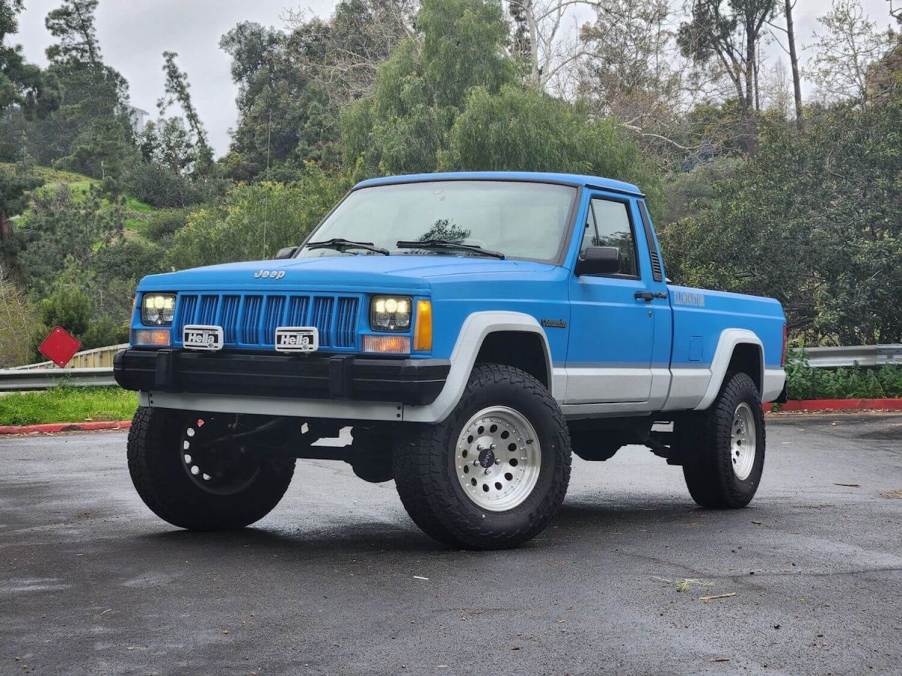 A blue 1990 Jeep Comanche Cherokee-based 4WD pickup truck in a parking lot for an auction photoshoot.