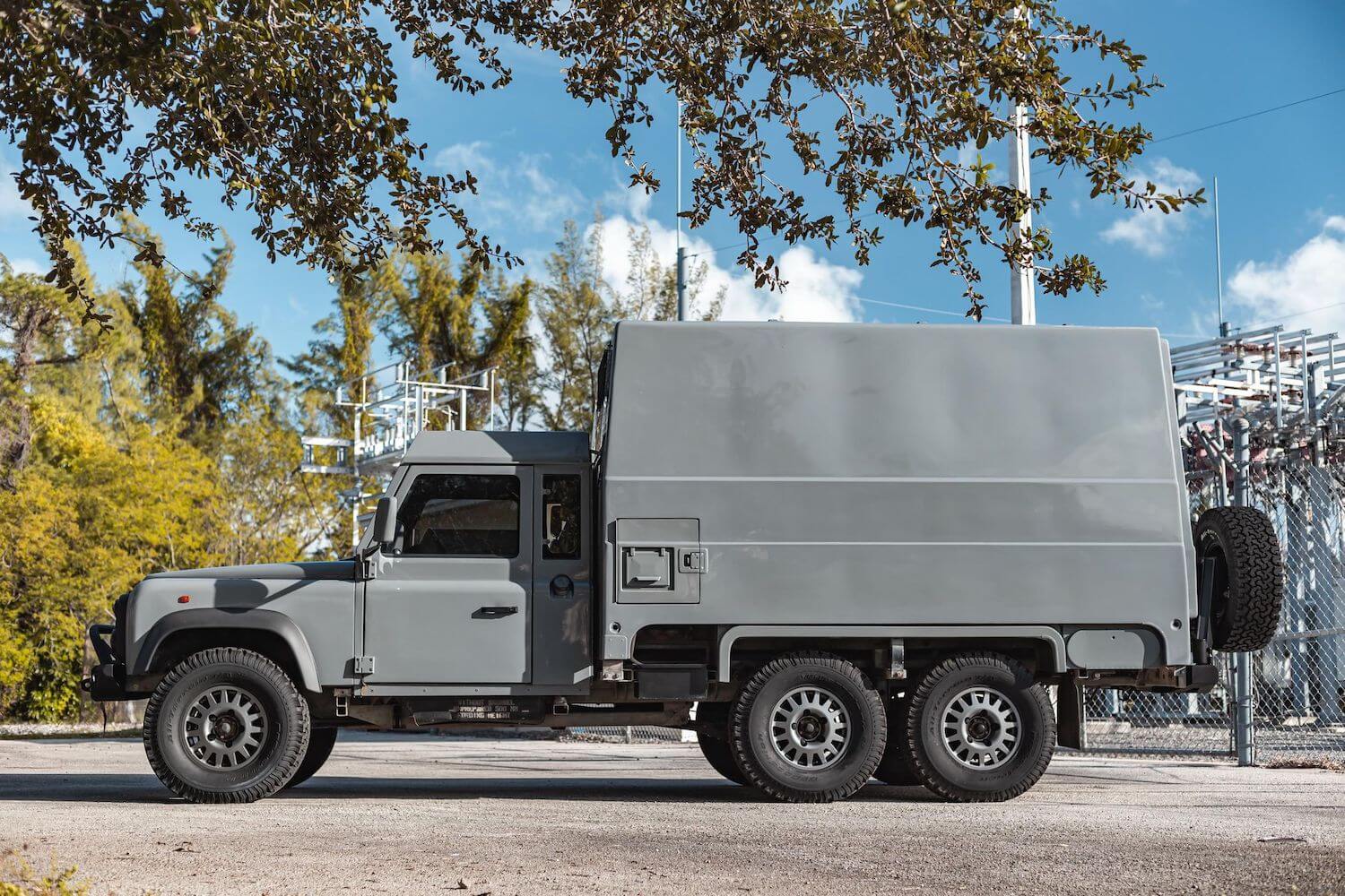 Profile photo of a Land Rover Defender 6x6 truck, all three of its axles clearly visible with trees in the background.