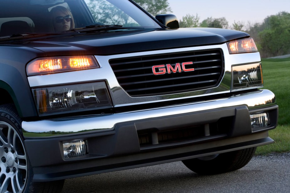 Closeup of the GMC logo in the grille of a compact Canyon pickup truck, trees visible in the background.