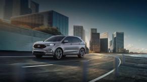 A dark gray 2023 Ford Edge midsize SUV model driving on a highway within an urban city. This Ford midsize SUV is not rated very highly.