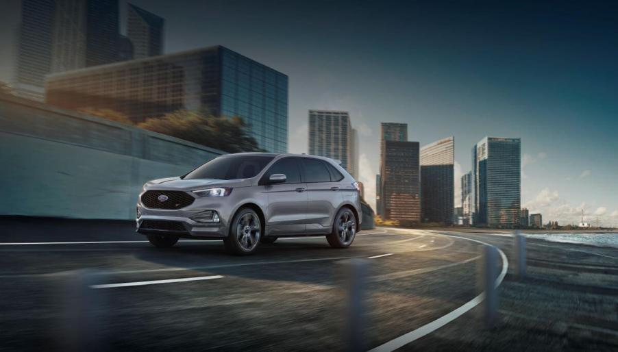 A dark gray 2023 Ford Edge midsize SUV model driving on a highway within an urban city. This Ford midsize SUV is not rated very highly.