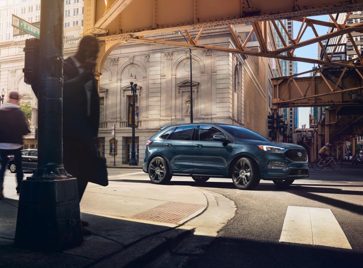 A blue 2023 Ford Edge midsize SUV model driving through a city crosswalk under metal girders