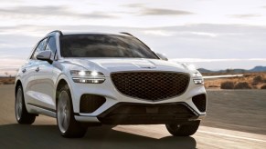 A white 2023 Genesis GV70 small luxury SUV is driving on the road.