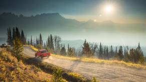 A red Jeep Gladiator Rubicon midsize pickup truck drives down a dirt road, mountains visible in the background.