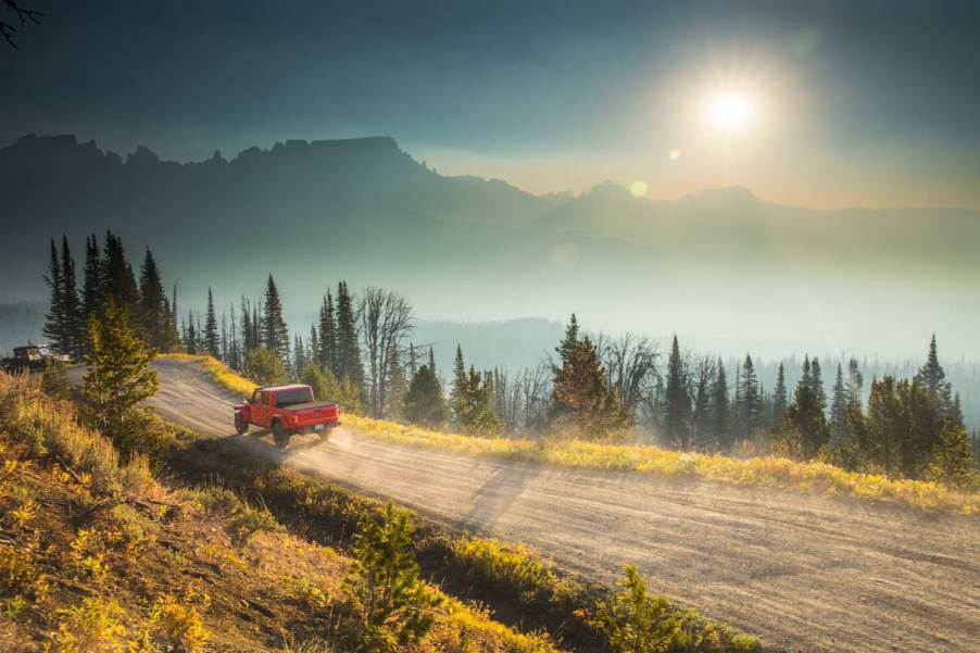 A red Jeep Gladiator Rubicon midsize pickup truck drives down a dirt road, mountains visible in the background.