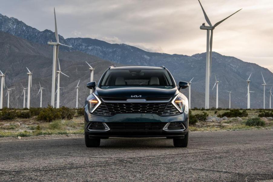 A blue-green 2023 Kia Sportage Hybrid compact SUV model parked in front of a field of windmills