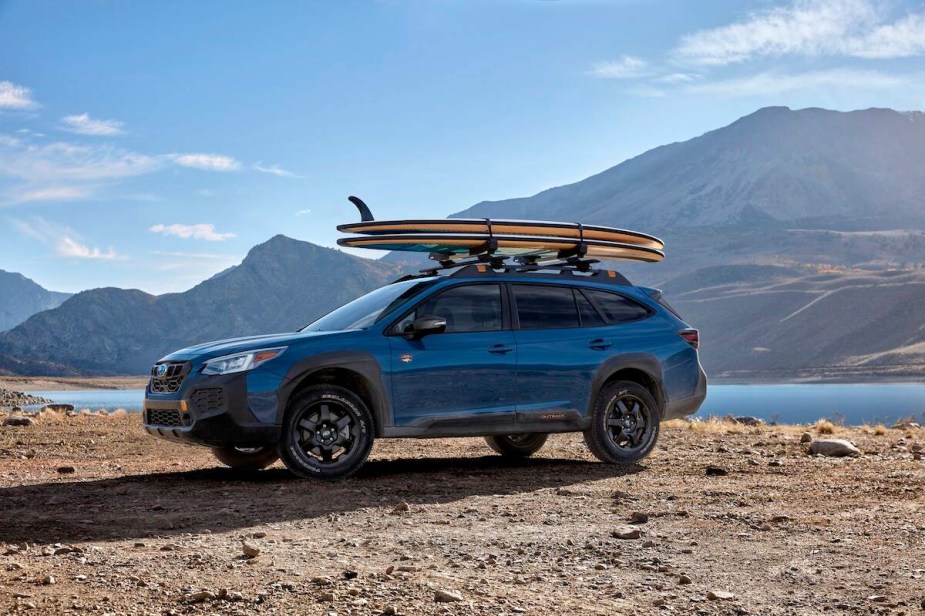 A blue 2023 Subaru Outback, which is a midsize SUV best for you, driving outdoors in front of a mountain and body of water. 