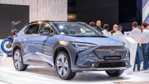 A blue 2023 Subaru Solterra EV SUV on display at an auto show with people around it.