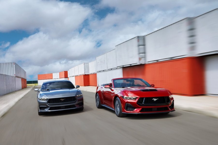 A set of new 2024 Ford Mustangs, including a GT, blast through a harbor showing off their gray-blue and red paint schemes.