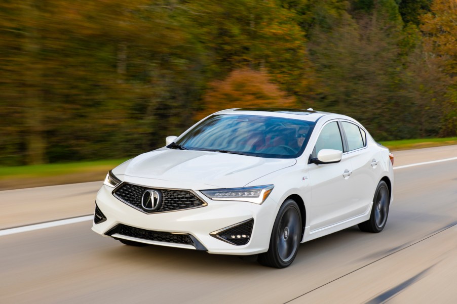 A white Acura ILX driving down the road in a wooded area.