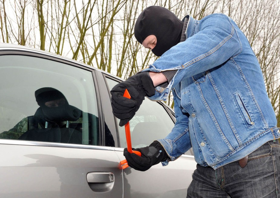 A man in a baklava and denim jacket uses a pry bar to get into a parked silver vehicle and steal it.