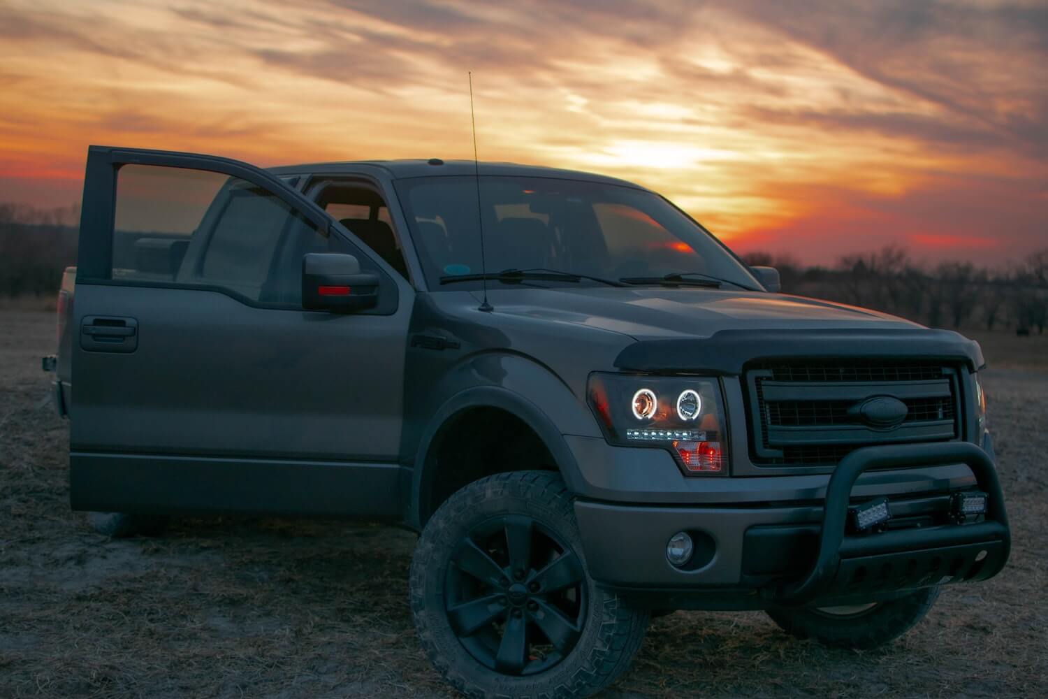 Gray Ford F-150 parked in front of a setting sun with its door open, trees visible in the background.