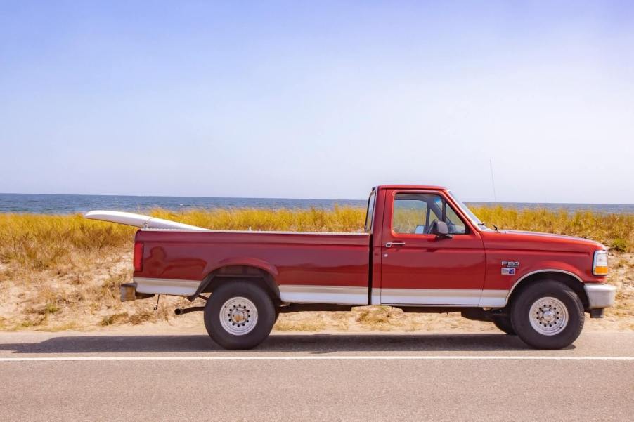 A regular cab Ford F-150 pickup truck parked by the water with a surfboard in its bed.