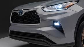 Front diagonal view of a grey 2023 Highlander Hybrid with headlamps and fog lamps illuminated. This model is one of the best Toyota hybrid models