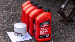 Five red quarts containers of oil ready to be poured into a motor during a change, alongside the oil to be replaced--all sitting on the pavement by a jack.