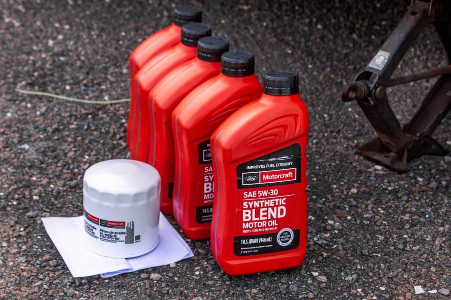 Five red quarts containers of oil ready to be poured into a motor during a change, alongside the oil to be replaced--all sitting on the pavement by a jack.