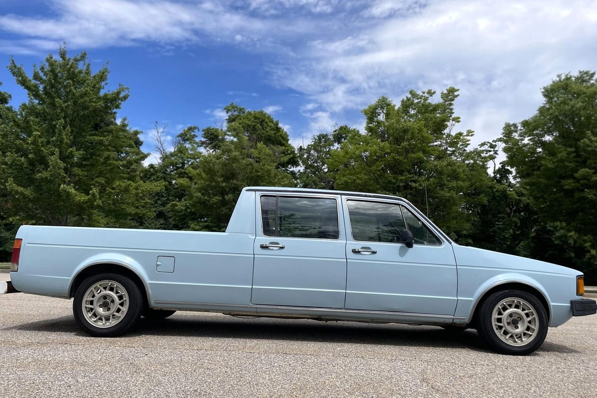 A blue 1981 Volkswagen Rabbit pickup that someone stretched into a sort of midsize VW truck, parked on pavement with trees in the background.