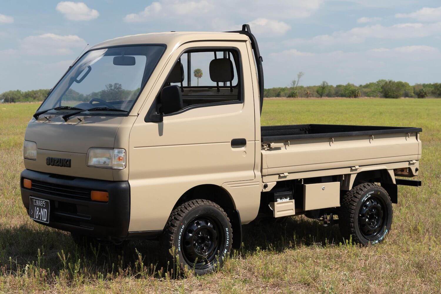 A tan Suzuki Carry "Kei-Class" mini truck parked in a field, trees visible in the background.