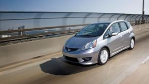 A silver-gray 2011 Honda Fit Sport hatchback model driving on a highway bridge overlooking an urban seaside city