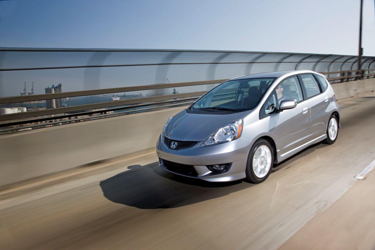 A silver-gray 2011 Honda Fit Sport hatchback model driving on a highway bridge overlooking an urban seaside city